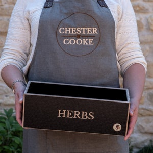 Könnte beinhalten: Eine rechteckige schwarze Kräuterbox aus Holz mit dem Wort "HERBS" in weißen Buchstaben. Die Box hat ein strukturiertes Muster und wird von einer Person gehalten, die eine graue Schürze mit dem Text "CHESTER COOKE" trägt.