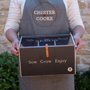 May include: A rectangular dark brown seed storage box with a clear lid, featuring a honeycomb pattern and the text "Sow Grow Enjoy" on the front. The box includes internal dividers labeled "Herbs," "Veg," and "Flowers," and a tan leather strap closure.