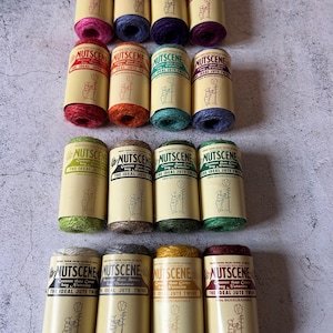 Puede incluir: Una colección de doce carretes de hilo de yute en varios colores, incluyendo rosa, azul, morado, rojo, naranja, turquesa, verde, marrón, amarillo y burdeos. Cada carrete está etiquetado con la marca "Nutscene" y presenta un dibujo de una persona con una planta.