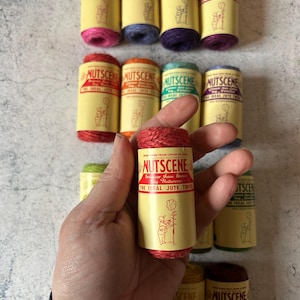 Puede incluir: Una colección de carretes de hilo de yute Nutscene en varios colores, como rosa, azul, morado, rojo, naranja y verde. Cada carrete tiene una etiqueta amarilla con el nombre de la marca, un gráfico y el texto "The Ideal Jute Twine".