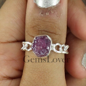 May include: A silver ring with a pink ruby gemstone set in a chain link band. The ring is worn on a person's thumb.