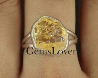 May include: A silver ring featuring a large, yellow, faceted gemstone. The gemstone is set in a simple, silver band. The ring is worn on a person's finger.
