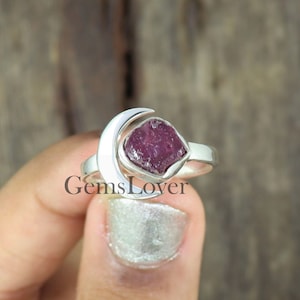May include: A silver ring featuring a crescent moon design and a raw ruby gemstone. The ring is displayed on a wooden surface.