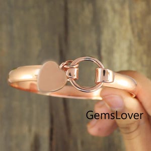 May include: A rose gold bangle bracelet featuring a smooth, polished finish. The bracelet includes a circular clasp mechanism attached to a small, solid heart-shaped charm. The metal has a warm, metallic pink hue throughout the entire piece.