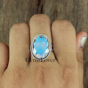 Peut inclure: Une bague en argent avec une grande pierre pr&eacute;cieuse ovale bleue. La pierre pr&eacute;cieuse a un effet arc-en-ciel iridescent. La bague a une bande en argent torsad&eacute;e.