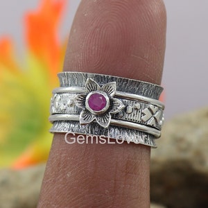 May include: A silver spinning ring featuring a pink gemstone set within a flower design. The ring has a textured finish and is adorned with small floral motifs.