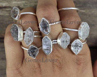 May include: A collection of silver rings, each featuring a unique, raw crystal gemstone. The gemstones vary in color, from clear to smoky gray, and are set in simple, elegant silver bands. The rings are displayed on a hand.