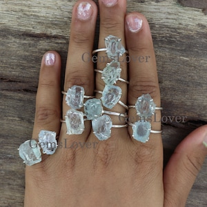 May include: A hand model wearing multiple silver rings with raw aquamarine gemstones. The rings are stacked on each finger. The rings are set with rough, natural aquamarine stones.