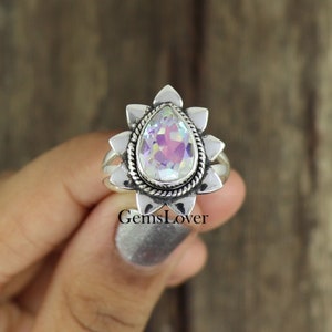 May include: A silver ring with a pear-shaped iridescent gemstone set in a floral design. The ring is on a finger.