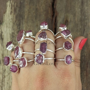 May include: A collection of silver rings featuring raw ruby gemstones. The rings are stacked on a hand, highlighting the unique shapes and colours of the rubies.
