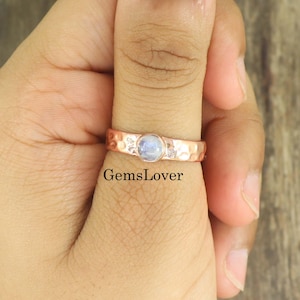 May include: A copper-toned ring featuring a central round, iridescent moonstone cabochon set in a bezel. The band has a hammered, textured finish with small, clear accent stones embedded on either side of the centre gem. The text GemsLover is visible.