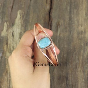 May include: A rose gold copper bracelet with a light blue gemstone set in a twisted wire design. The bracelet is open and adjustable.