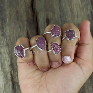 May include: Five silver rings with pink, rough-cut gemstones. The rings are worn on a hand.