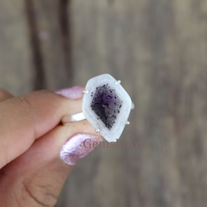 May include: A silver ring with a distinctive, geometric gemstone. The stone has a white outer edge and a dark purple and black centre. The ring is held by four silver prongs.