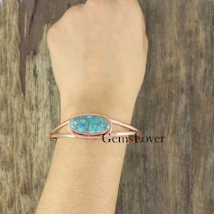 Peut inclure: Un bracelet manchette en cuivre avec une pierre turquoise sertie au centre. La pierre présente un motif bleu et vert marbré. Le bracelet est ouvert et réglable.