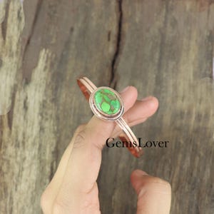 May include: A copper bangle bracelet with a green oval turquoise stone set in a silver bezel. The bracelet is open and adjustable.