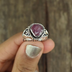 May include: A silver ring featuring a pink gemstone set in a twisted silver band. The ring has a decorative design on the sides.