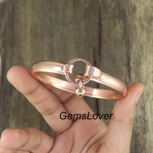 May include: A rose gold bracelet with a circular clasp and a small dangling sphere. The bracelet is held in a hand against a wooden background. The text "GemsLover" is visible on the image.