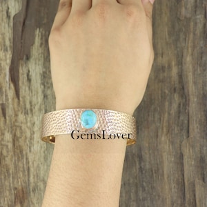 May include: A silver cuff bracelet with a hammered texture and a turquoise stone set in the center. The bracelet is on a person's wrist.