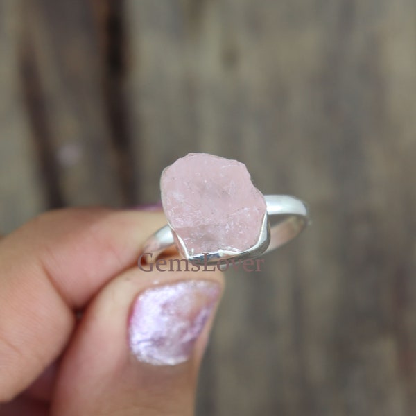 May include: A silver ring featuring a raw, light pink rose quartz gemstone. The stone has a rough, natural texture and is set on a simple silver band. The ring is displayed against a blurred wooden background.