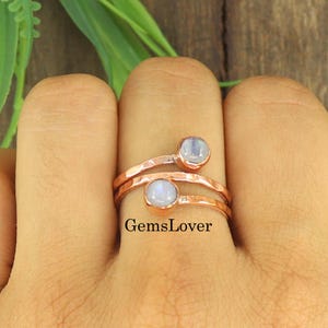 Peut inclure: Une bague ouverte en fil de cuivre martelé, ornée de deux cabochons ronds en pierre de lune blanche translucide sertis dans des chatons simples. L'anneau métallique présente une finition texturée et rustique avec le texte GemsLover inscrit.