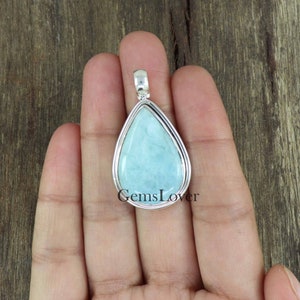 May include: A teardrop-shaped pendant with a light blue gemstone set in a silver frame. The pendant is held in a hand, with the text "GemsLover" visible below the pendant.