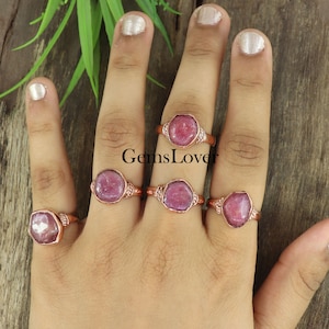 Op de afbeelding: Vijf koperen ringen met roze edelsteen bezettingen. De ringen zijn op een hand, met de edelstenen naar de camera gericht. De tekst "GemsLover" is zichtbaar op de achtergrond.