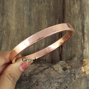Könnte beinhalten: Ein schlichtes Kupfer-Armreif-Armband. Das Armband ist glatt und glänzend, mit einer leicht abgerundeten Kante. Es ist ein klassisches und zeitloses Design.
