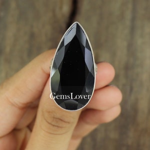 May include: A large, teardrop-shaped black gemstone ring with a silver band. The faceted stone reflects light, and the ring is held by a hand. The text "GemsLover" is visible on the ring.