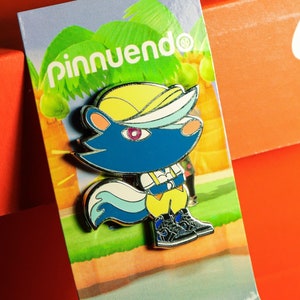 May include: A colourful enamel pin featuring a cartoon animal wearing a yellow hat and blue shirt. The pin is on a card with the text "pinnuendo" in green letters.