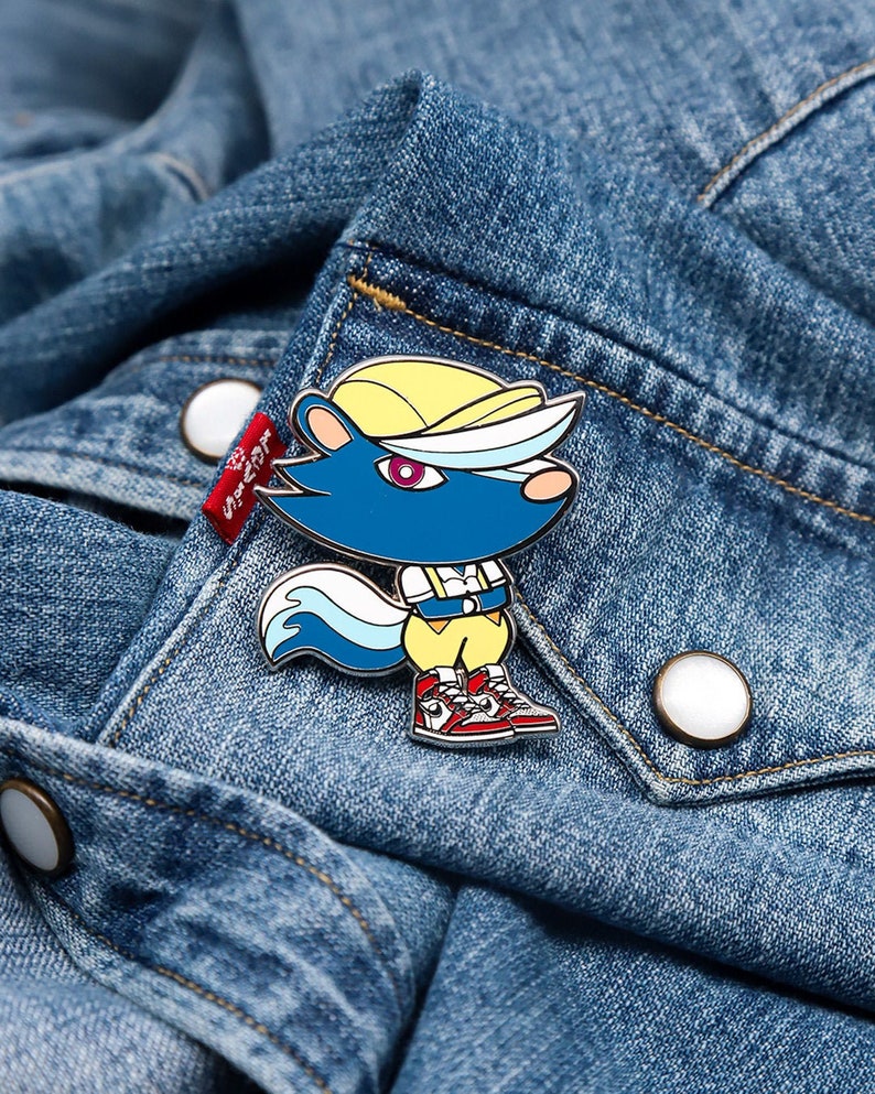 May include: A blue and yellow enamel pin featuring a cartoon character with a white and blue tail, wearing a yellow shirt, red shoes, and a yellow hat. The pin is on a blue denim jacket.
