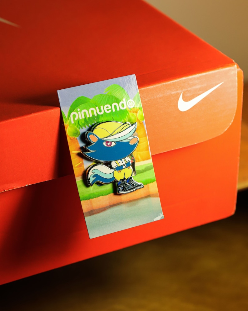 May include: A colorful enamel pin featuring a cartoon animal wearing a yellow hat and blue shirt. The pin is on a card with the text "pinnuendo" in green letters.