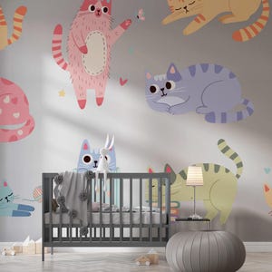 Pode incluir: Uma parede cinza de ber&ccedil;&aacute;rio com ilustra&ccedil;&otilde;es coloridas de gatos de desenho animado. Os gatos est&atilde;o em v&aacute;rias poses e cores, incluindo rosa, azul e amarelo. Um ber&ccedil;o cinza com um cobertor e uma pequena l&acirc;mpada est&atilde;o em primeiro plano.