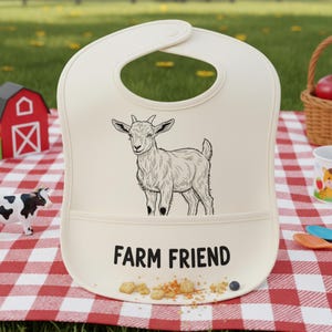 May include: Cream baby bib featuring a black and white goat illustration and the text "FARM FRIEND". The bib has a pocket and is on a red and white checked picnic blanket. Other farm-themed toys are nearby.