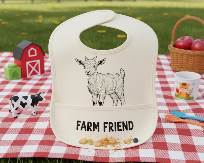 May include: Cream baby bib featuring a black and white goat illustration and the text "FARM FRIEND". The bib has a pocket and is on a red and white checked picnic blanket. Other farm-themed toys are nearby.