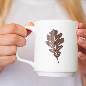 May include: A white ceramic mug with a brown oak leaf design. The mug is being held by a person's hands.