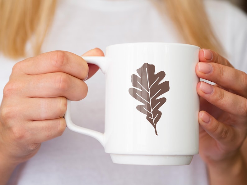 May include: A white ceramic mug with a brown oak leaf design. The mug is being held by a person's hands.