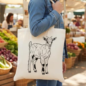 May include: A cream tote bag with a black and white goat illustration. The goat is depicted standing, with detailed fur and horns. The bag is held in front of a farmer's market, with fresh produce visible in the background.