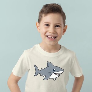 May include: A cream-colored t-shirt featuring a cartoon shark illustration. The shark is gray with white accents and a smiling expression. The shirt is worn by a young person, smiling at the camera, against a light blue background.