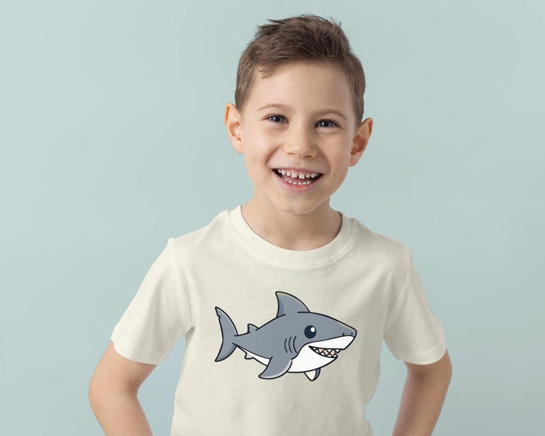 May include: A cream-colored t-shirt featuring a cartoon shark illustration. The shark is gray with white accents and a smiling expression. The shirt is worn by a young person, smiling at the camera, against a light blue background.