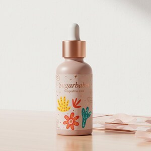 May include: A light pink bottle with a dropper top and a rose gold cap. The bottle has a label with the text "Sugarbabs Temptation Line" and colorful floral illustrations. The bottle is on a wooden surface.