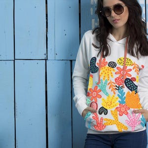 May include: White hoodie with a colorful floral design. The design features orange, yellow, and blue flowers and abstract shapes. The hoodie has a drawstring and a front pocket. The model is wearing blue jeans.