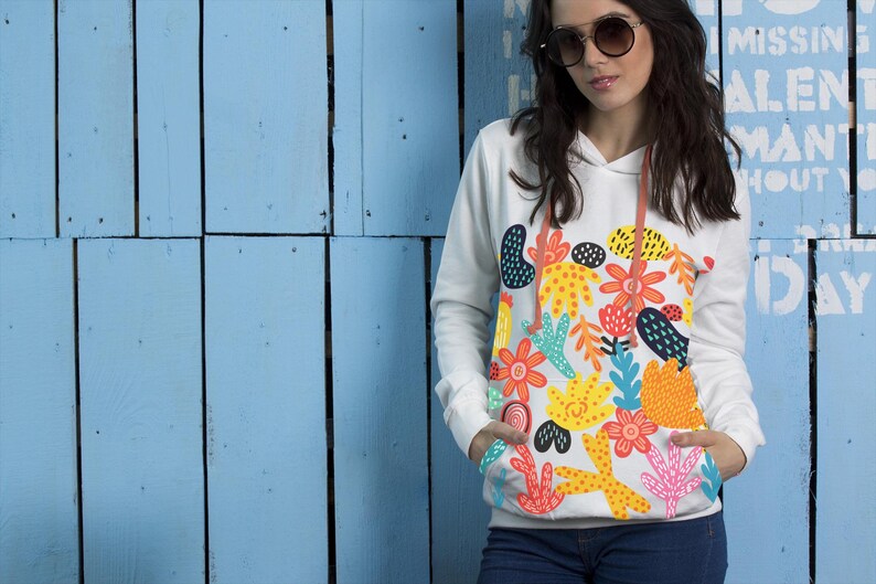 May include: White hoodie with a colorful floral design. The design features orange, yellow, and blue flowers and abstract shapes. The hoodie has a drawstring and a front pocket. The model is wearing blue jeans.