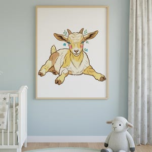 May include: A framed art print of a cartoon goat in yellow, brown, and white, with small green floral accents. The goat is lying down, facing forward. The artwork is displayed in a light-coloured wooden frame, suitable for a nursery or child's room.