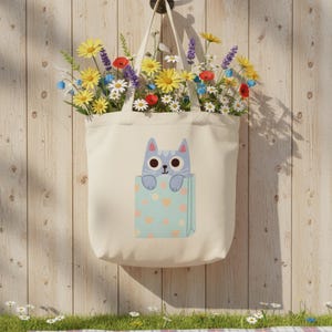 Pode incluir: Uma sacola creme com um desenho de gato em um design de bolso, cheia de flores coloridas. O gato &eacute; azul claro com olhos grandes. O bolso &eacute; azul claro com cora&ccedil;&otilde;es rosa. A sacola est&aacute; pendurada em uma parede de madeira.