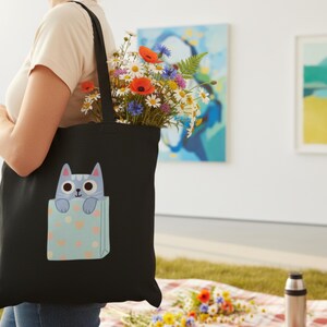 Pode incluir: Bolsa tote preta com um design de gato de desenho animado, cheia de um vibrante buqu&ecirc; de flores. O gato azul claro com olhos grandes espreita de um bolso azul claro decorado com cora&ccedil;&otilde;es rosa. Um acess&oacute;rio elegante e pr&aacute;tico.