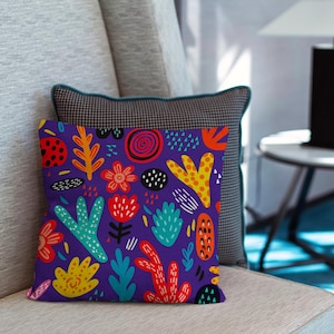 May include: A decorative throw pillow with a vibrant, colorful floral pattern. The pillow features a deep purple background with various shapes in red, yellow, orange, and teal. The pillow is placed on a light gray sofa, with a patterned pillow behind it.