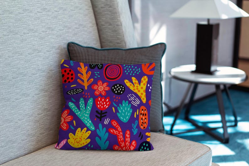 May include: A decorative throw pillow with a vibrant, colorful floral pattern. The pillow features a deep purple background with various shapes in red, yellow, orange, and teal. The pillow is placed on a light gray sofa, with a patterned pillow behind it.