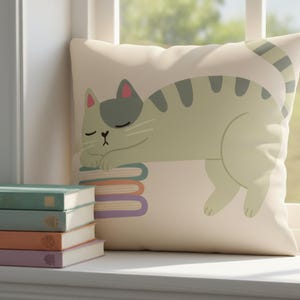 Pode incluir: Uma almofada decorativa com uma ilustra&ccedil;&atilde;o de um gato a dormir em tons de verde, cinza e rosa. O gato est&aacute; deitado numa pilha de livros com lombadas coloridas. A almofada est&aacute; colocada numa janela branca, ao lado de uma pilha de livros.