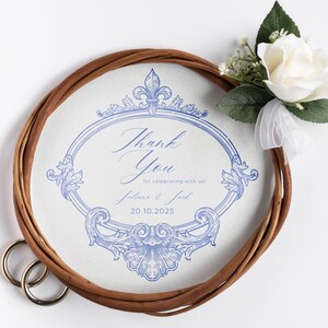 Puede incluir: Un recuerdo de boda con un dise&ntilde;o floral azul y la frase "Thank You". Una corona trenzada marr&oacute;n rodea el dise&ntilde;o, con una rosa blanca y vegetaci&oacute;n adjunta. Se ven dos anillos dorados.